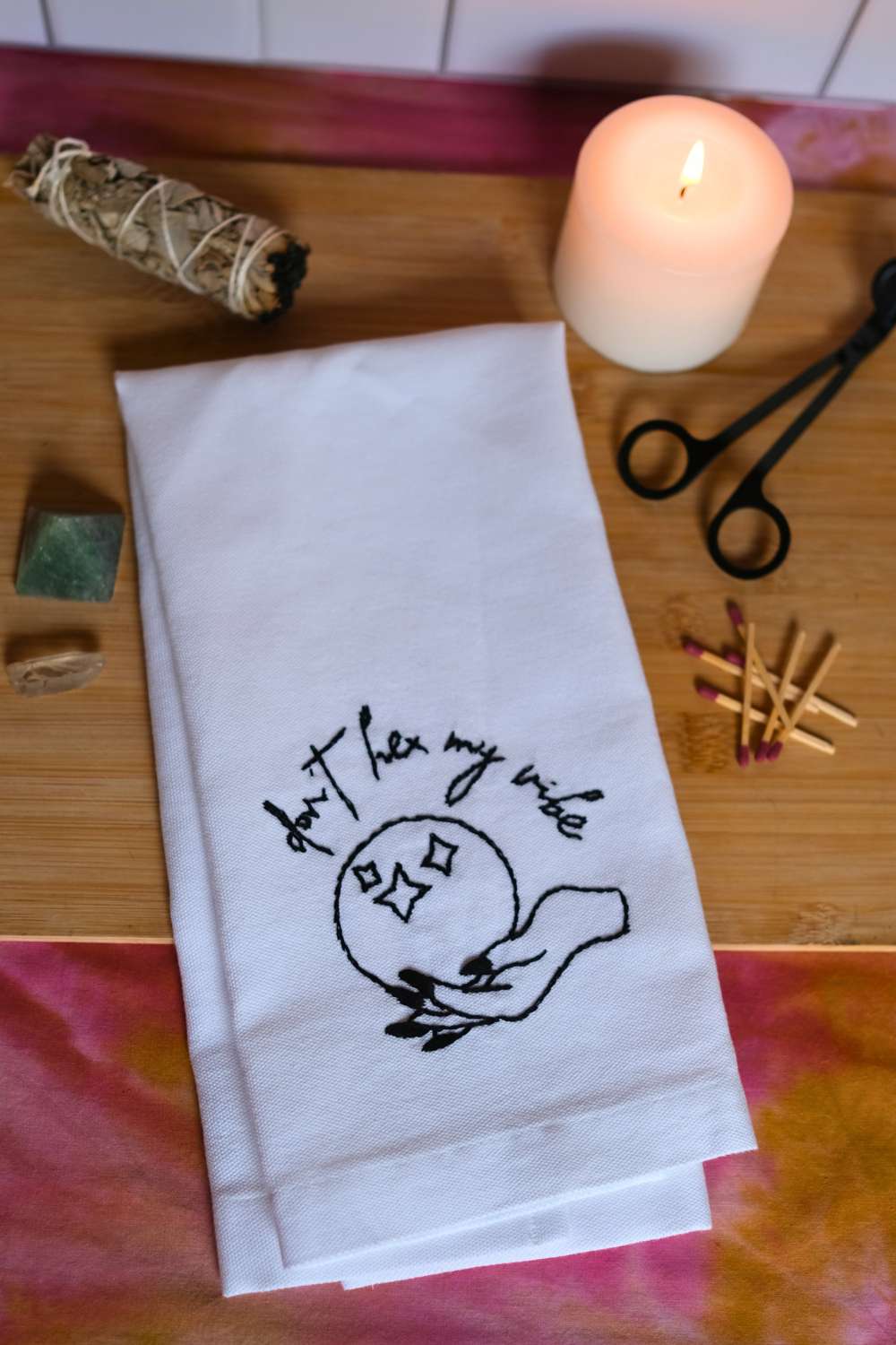 White tea towel with black DIY Halloween hand embroidery reads "don't hex my vibe" in cursive font and shows a witch hand holding a crystal ball. Towel sits atop a wood cutting board surrounded by sage, crystals, lit white candle, black wick trimmer, and matches.