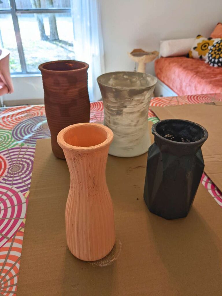 Pink, gray, white, and brown vases sit on top of a piece of cardboard above a colorful tablecloth. The vases are shown mid-project with a light coat of paint on them.