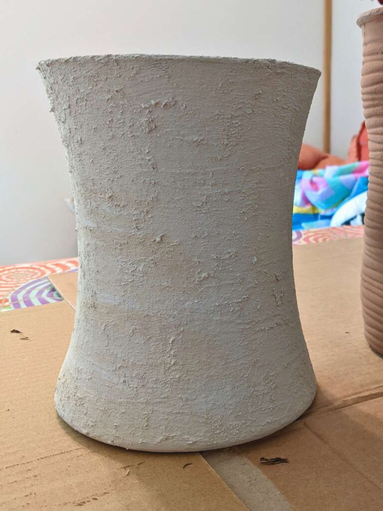 An empty white vase with grainy texture sits on cardboard.