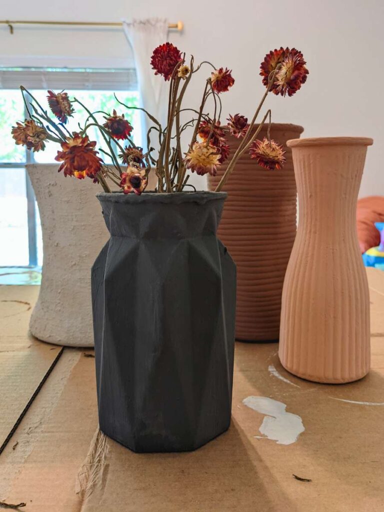 Four vases rest on a piece of cardboard. A dark gray vase in the foreground holds dried pink flowers. A pink, brown, and white vase sit empty in the background.