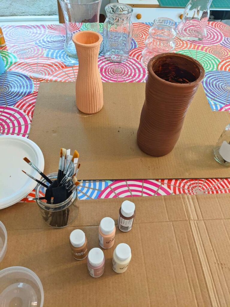 A tabletop is adorned with a brightly colored table cloth, cardboard, paint tubes, paintbrushes, and vases.