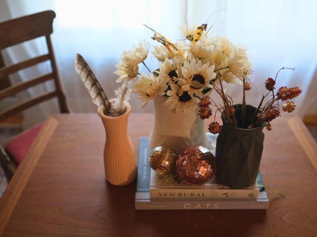 Gray, white, and pink vases filled with dried flowers and feathers sit beside two copper disco balls atop two books on a dining room table.