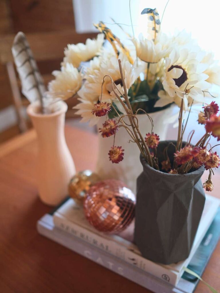 Gray, white, and pink vases filled with dried flowers and feathers sit beside two copper disco balls atop two books on a dining room table.
