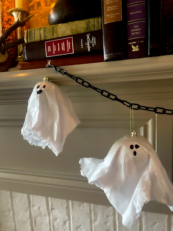 Budget-friendly DIY Halloween decor: Two white tissue paper ghosts hang from a black chain garland on a white fireplace mantle. On top of the mantle are books and candles. 