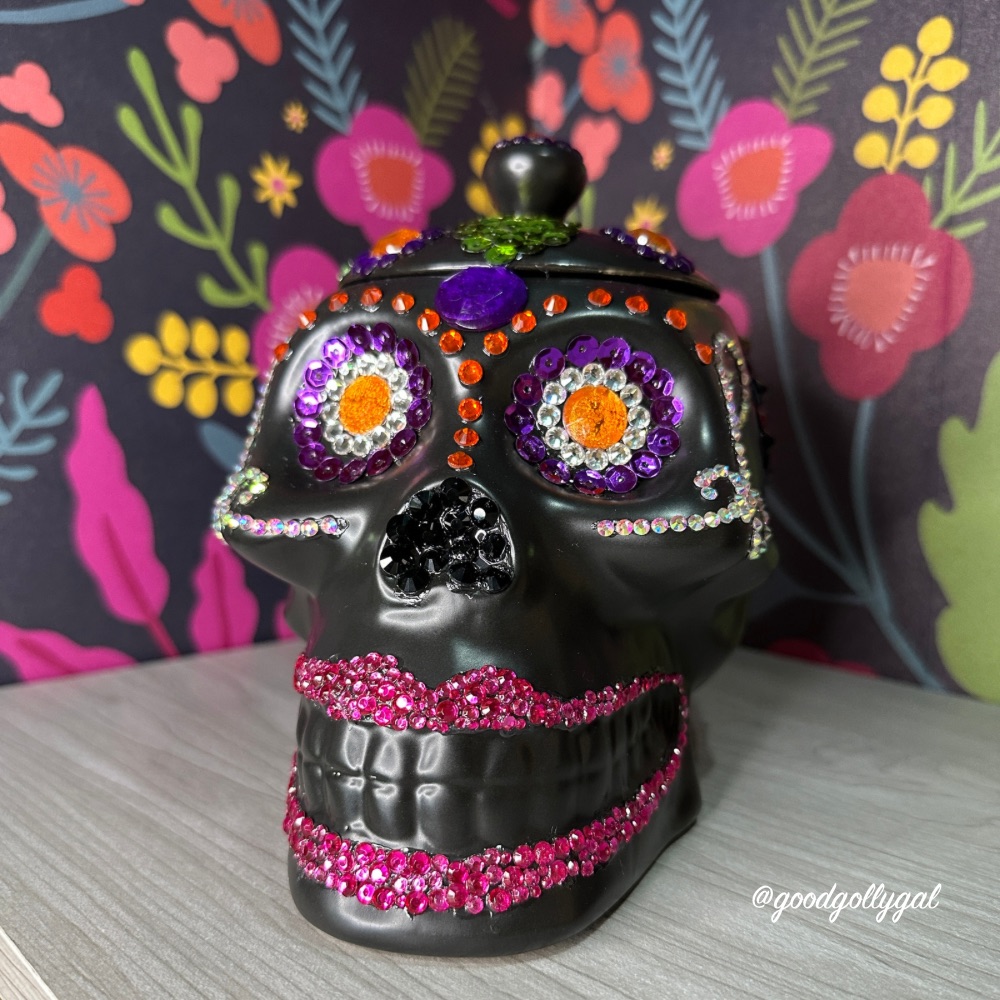 Budget-friendly DIY Halloween decor: Black skull-shaped cookie jar bedazzled with rhinestones sits on a wooden shelf in front of a colorful floral painted mural. 
