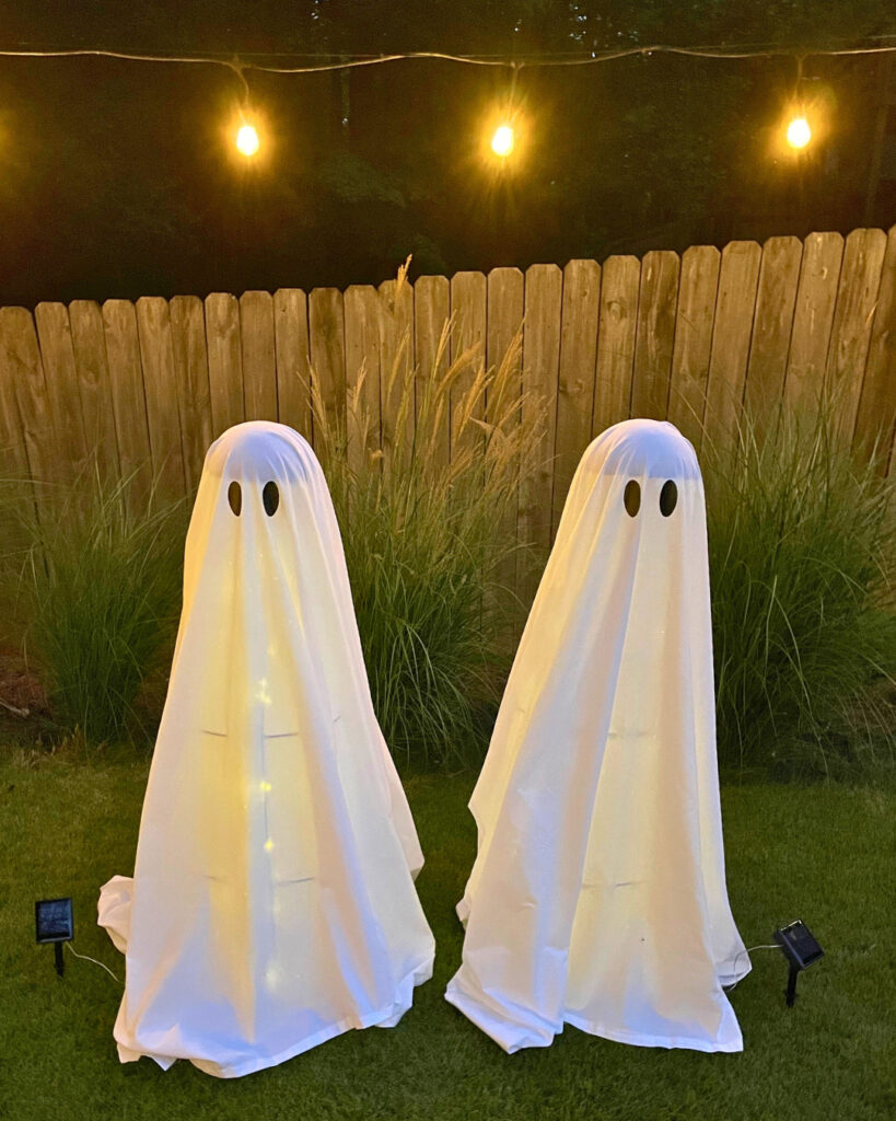 Budget-friendly DIY Halloween decor: Two white ghosts on a green lawn lit up with solar lights in front of greenery and wooden fence. 